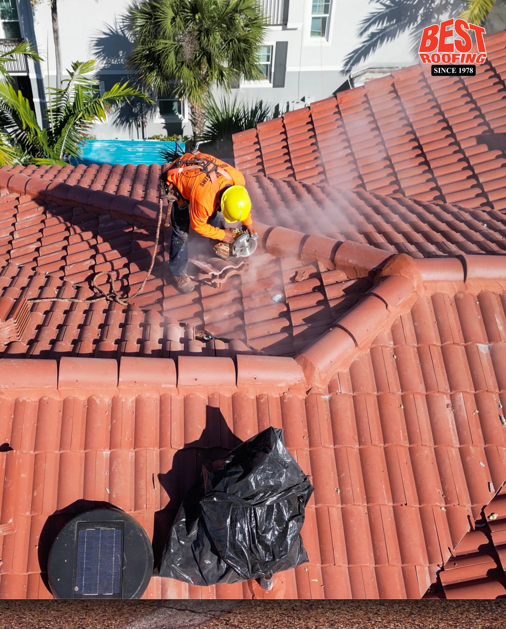 Worker in safety gear cutting red clay roof tiles on a sunny day with palm trees and pool in background. 